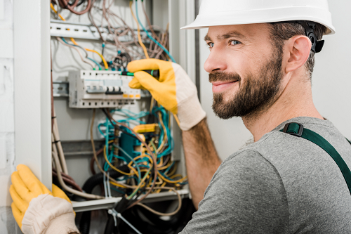 Smiling Electrician wearing white safety hat and yellow gloves while inspecting a electrical panel