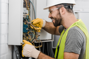 Smiling Technician wearing a Yellow safety Jacket, a grey tshirt, white safety hat and gloves inspecting an electrical panel